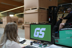 Frau im Lager am Computer, Gabelstapler im Hintergrund.
