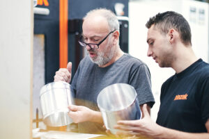 Zwei Männer untersuchen Metallobjekte in Werkstatt.